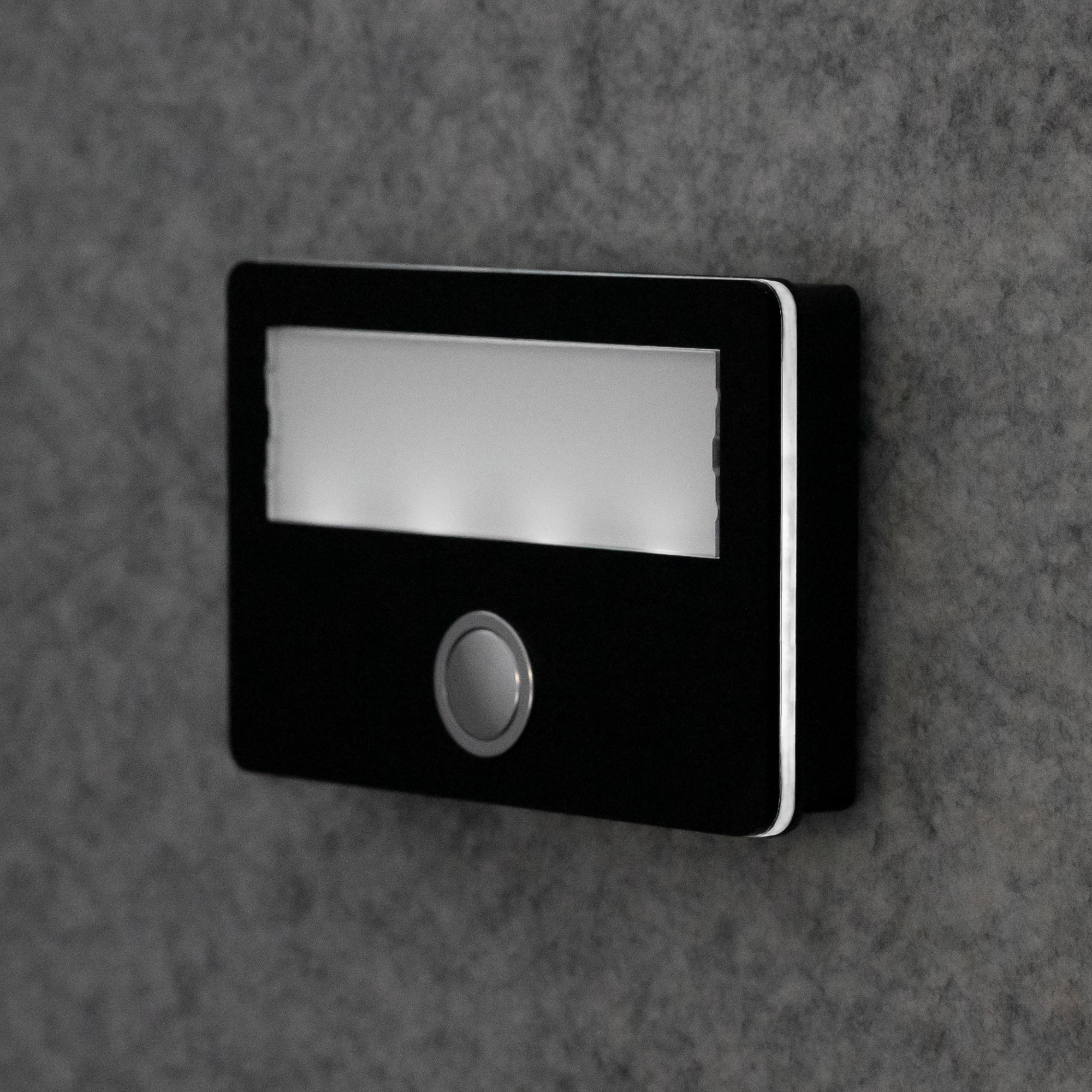 Seitlich beleuchtete Etagenplatte mit rundem Taster und Namensschild, Ansicht von schräg vorne rechts an der Wand.