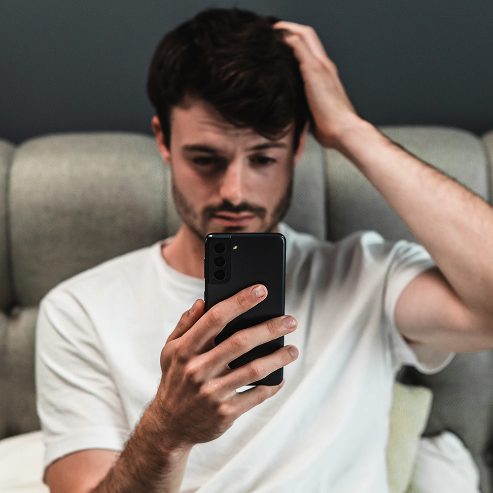 Mann sitzt im Bett, hält sein Smartphone in der Hand und fasst sich an den Kopf