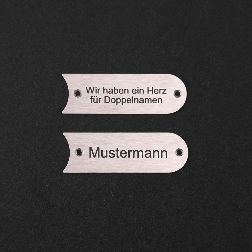 Zwei Edelstahl-Gravierschilder untereinander; auf dem unteren steht "Mustermann", auf dem oberen steht "Wir haben ein Herz für Doppelnamen"