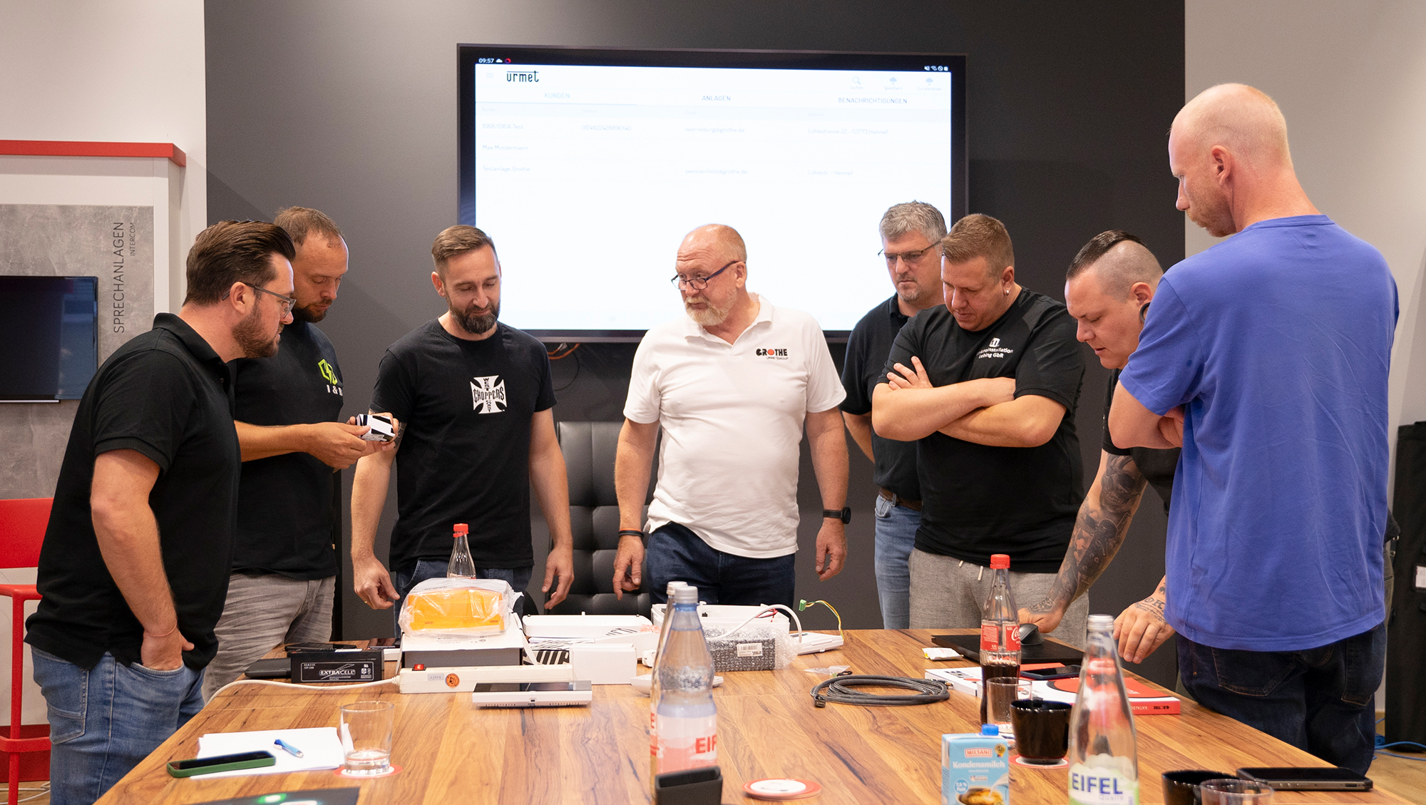 Mehrere Männer stehen um den Tisch und Showroom und blicken auf den Tisch; im Hintergrund ist ein großer Monitor zu sehen