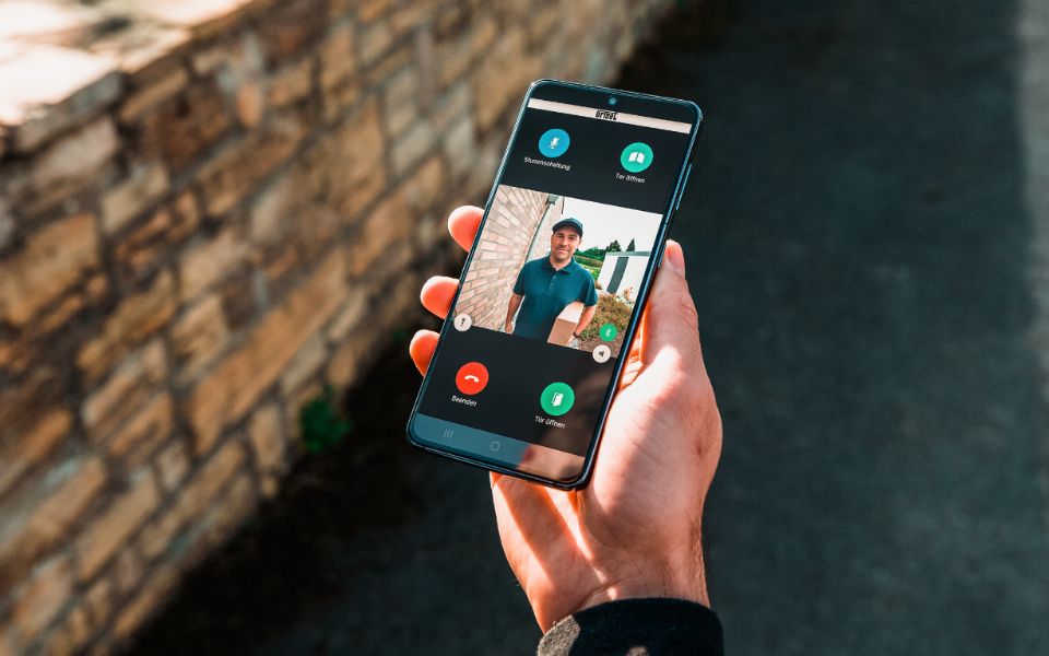 Hand hält Smartphone; auf dem Display ist die App zu erkennen, die einen Paketboten vor der Tür zeigt