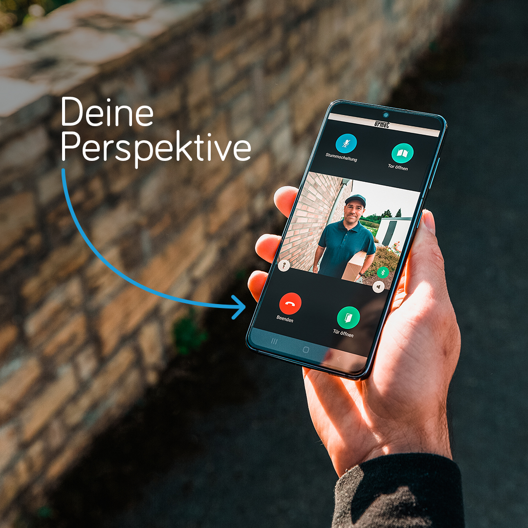 Hand hält Smartphone; auf dem Display ist die App zu erkennen, die einen Paketboten vor der Tür zeigt; daneben steht an einem Pfeil "Deine Perspektive"