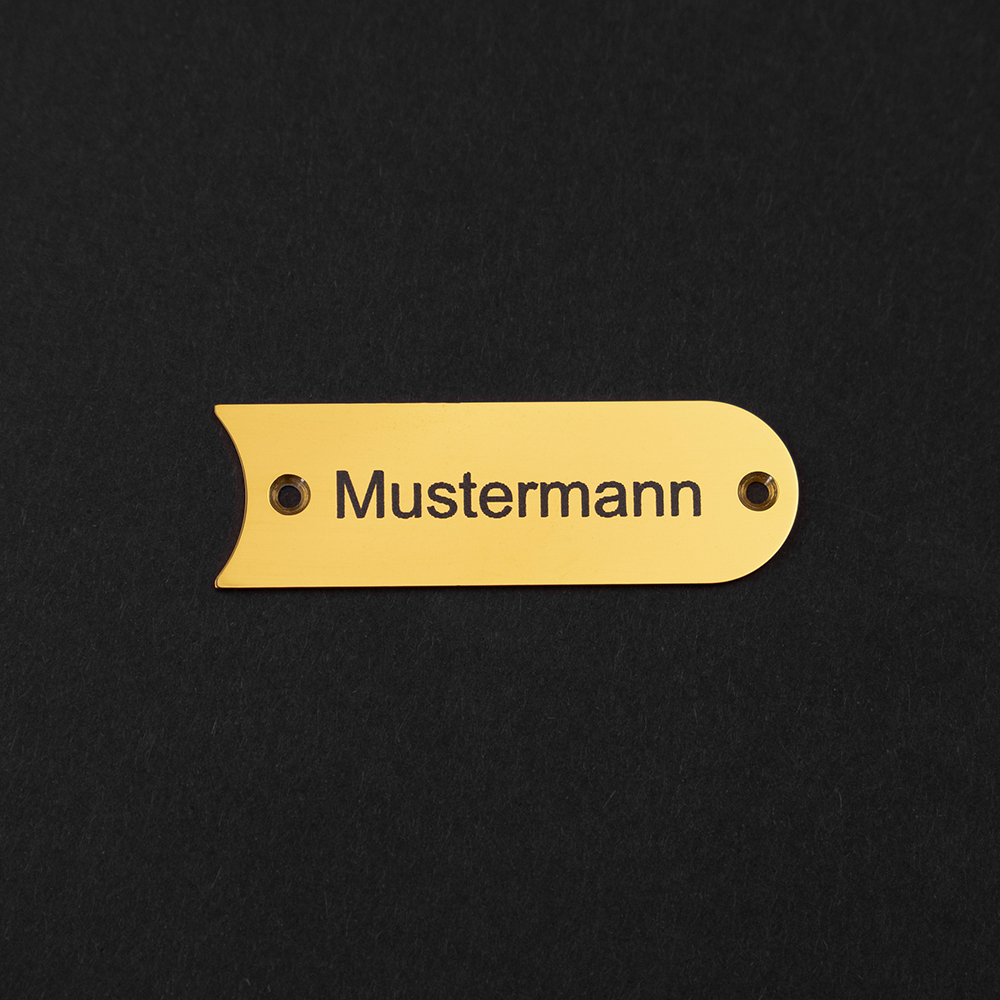 EIn messingfarbenes Gravierschild liegt auf schwarzem Grund; auf dem Schild steht "Mustermann"