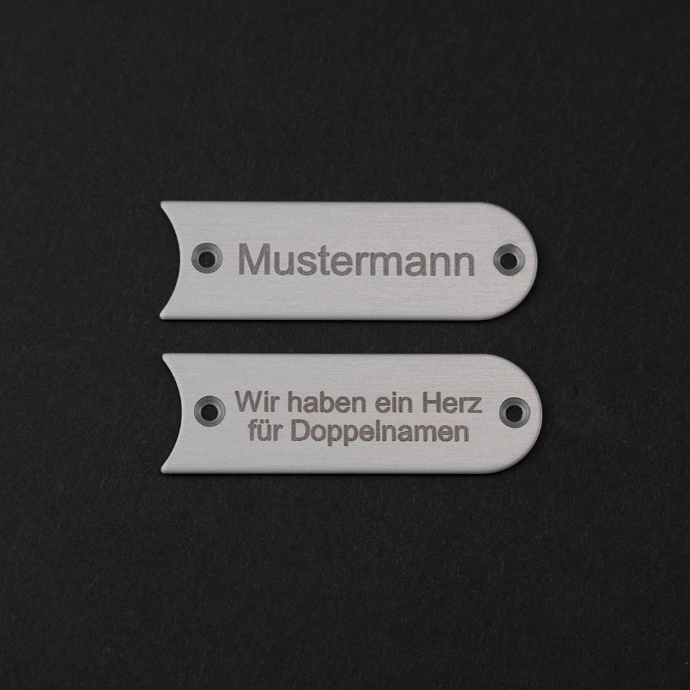 Zwei Edelstahl-Gravierschilder untereinander; auf dem oberen steht "Mustermann", auf dem unteren steht "Wir haben ein Herz für Doppelnamen"