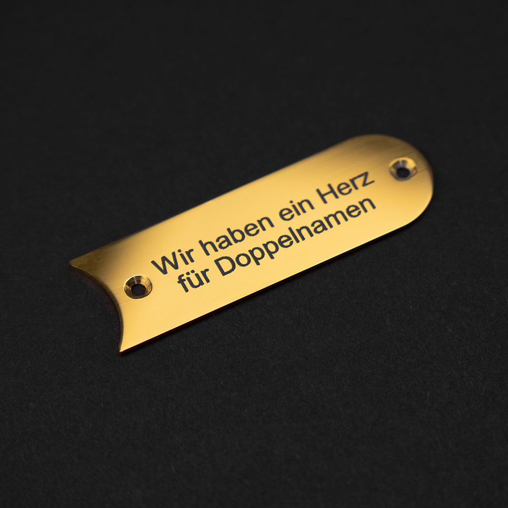 EIn messingfarbenes Gravierschild liegt auf schwarzem Grund; auf dem Schild steht "Wir haben ein Herz für Doppelnamen"