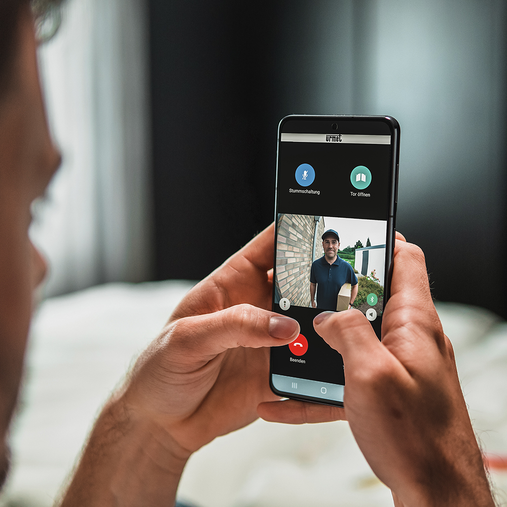 Blick über die Schulter eines Mannes, der auf sein Smartphone blickt; auf dem Display ist die App zu erkennen, die einen Paketboten vor der Tür zeigt