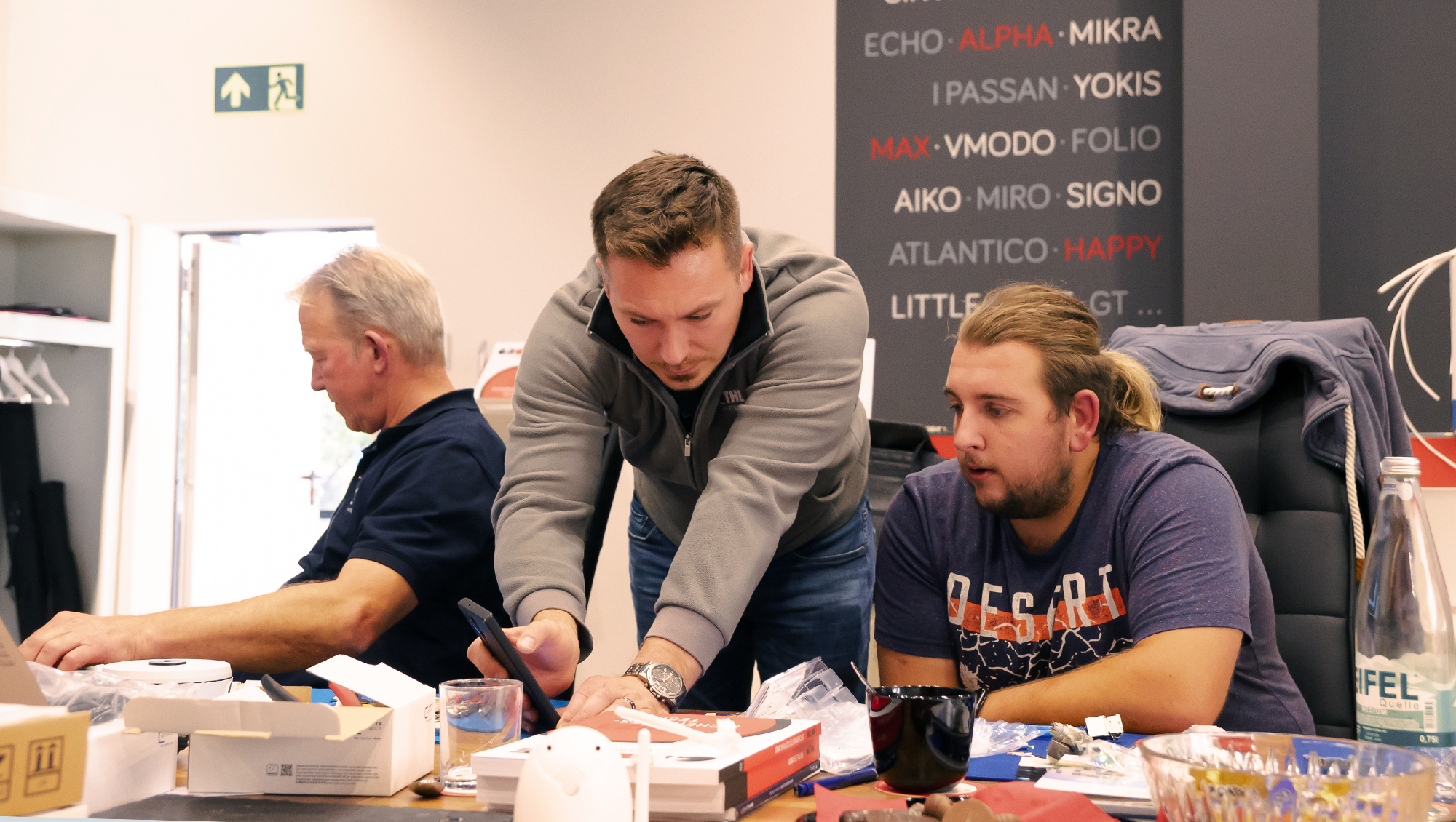 Am Tisch im Showroom sitzen zwei Schulungsteilnehmer; dazwischen beugt sich ein Grothe-Mitarbeiter vor und zeigt etwas auf seinem Smartphone