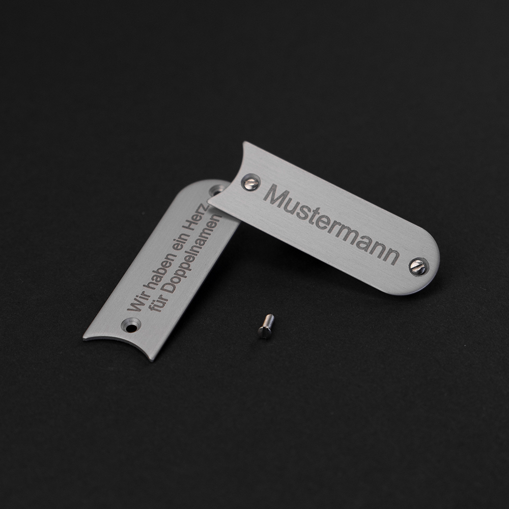 Zwei Edelstahl-Gravierschilder liegen halb übereinander; auf dem oberen steht "Mustermann", auf dem unteren steht "Wir haben ein Herz für Doppelnamen"
