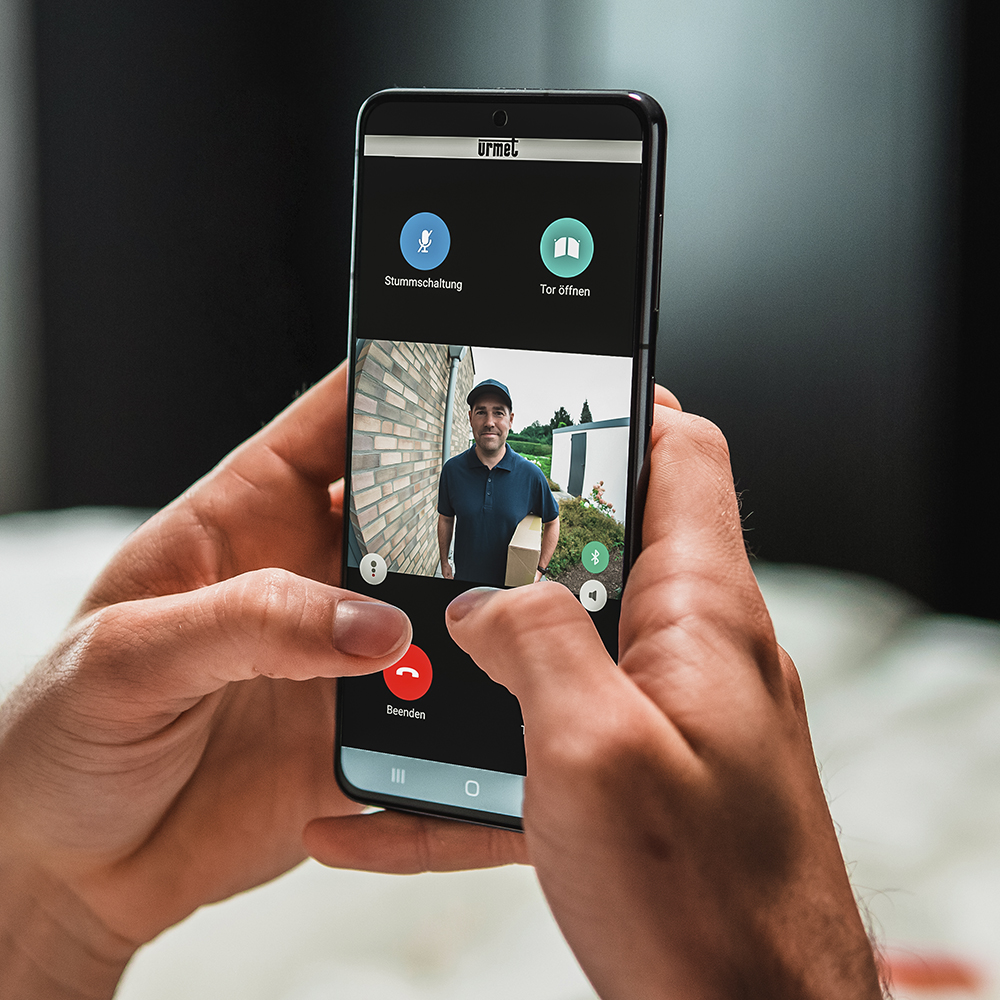 Blick über die Schulter eines Mannes, der auf sein Smartphone blickt; auf dem Display ist die App zu erkennen, die einen Paketboten vor der Tür zeigt