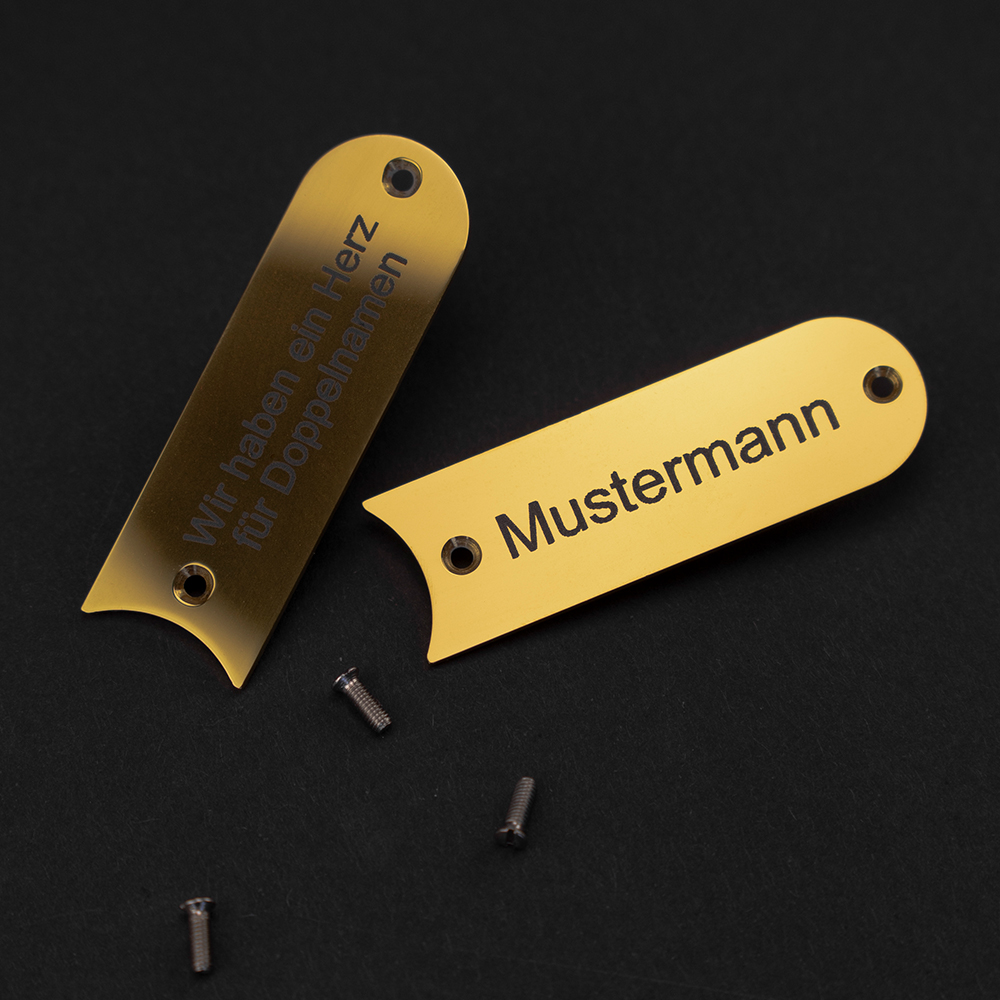 Zwei messingfarbene Gravierschilder liegen untereinander auf schwarzem Grund; auf dem vorderen steht "Mustermann", das hintere liegt im Halbschatten