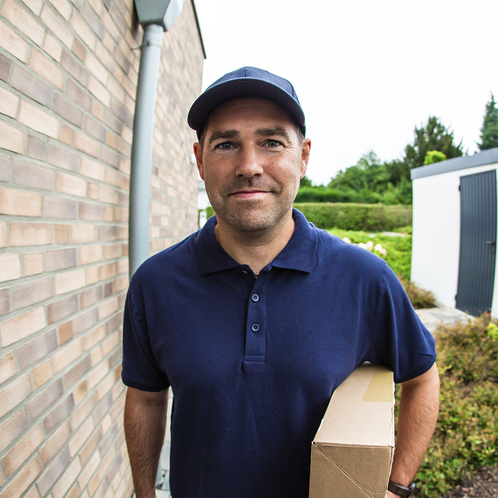 Kamerabild der vidoora: Vor der Tür steht ein Paketbote mit blauem T-Shirt und blauer Kappe, der unter dem Arm ein Paket hält und freundlich in die Kamera blickt