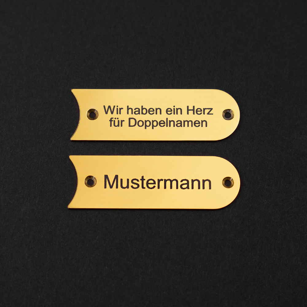 Zwei messingfarbene Gravierschilder liegen untereinander auf schwarzem Grund; auf dem unteren steht "Mustermann", auf dem oberen steht "Wir haben ein Herz für Doppelnamen"