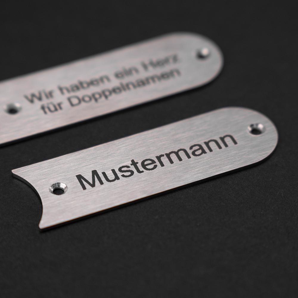 Zwei Edelstahl-Gravierschilder untereinander; auf dem unteren steht "Mustermann", auf dem oberen steht "Wir haben ein Herz für Doppelnamen"