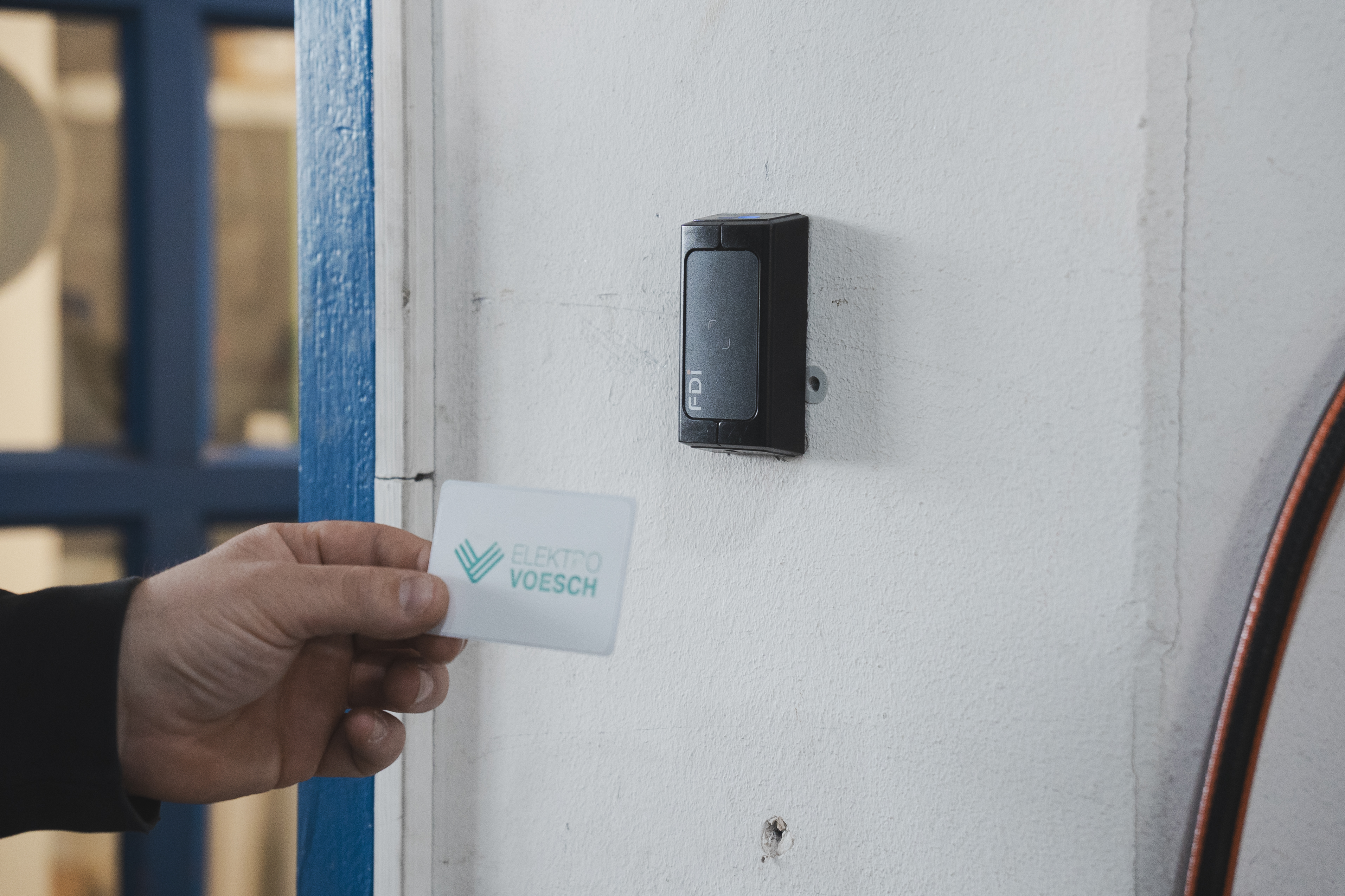 Lesegerät an einer weißen Wand; eine Hand hält eine Näherungskarte mit dem Aufdruck "Elektro Voesch" davor