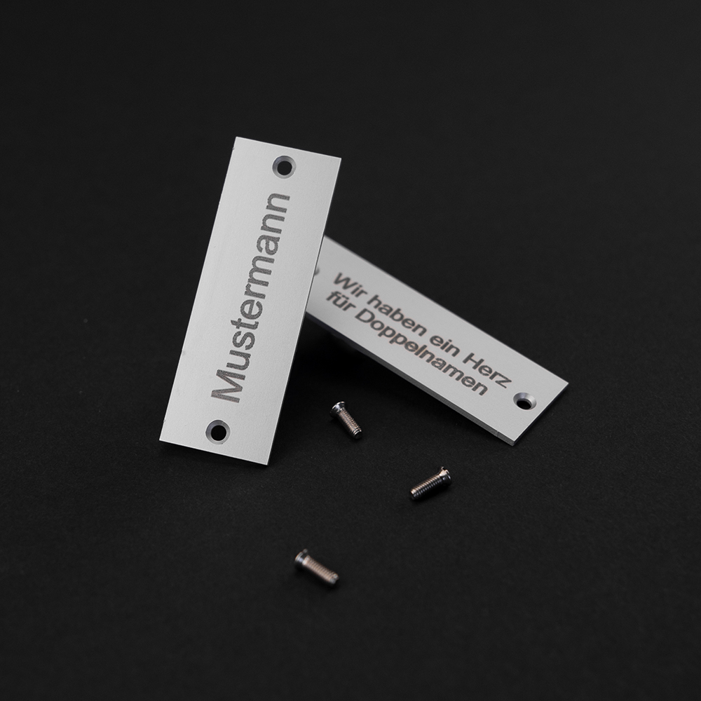 Zwei Edelstahl-Gravierschilder liegen halb übereinander; auf dem oberen steht "Mustermann", auf dem unteren steht "Wir haben ein Herz für Doppelnamen"